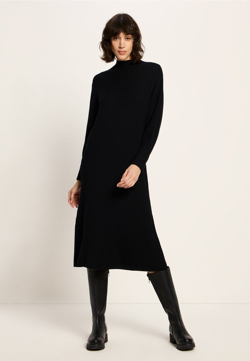 Schwarzes geripptes Strickkleid mit langen Ärmeln, hohem Kragen und Midi-Länge. Getragen mit knielangen schwarzen Stiefeln für einen kompletten Look.