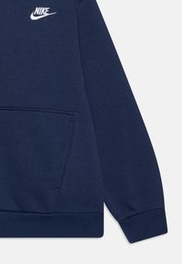 Tamsiai mėlynas Nike džemperis su kengūros kišenėle, su raišteliais rankogaliuose ir apačioje, pasižymintis mažu baltu logotipu ant krūtinės. Minkšta tekstūra.