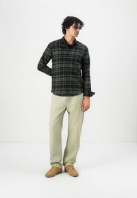 Camisa de cuadros negra y verde, jeans verde claro y zapatillas de deporte beige. Las gafas de sol completan el conjunto. Se nota el cabello rizado.