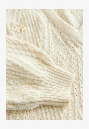 Pull en tricot crème avec un col et des poignets côtelés, présentant un design texturé et un accent délicat de nœud en dentelle.