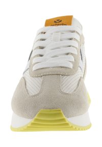 Baskets blanches et grises avec un embout en daim, des panneaux latéraux en mesh, une semelle extérieure jaune et une étiquette de langue orange présentant un logo.