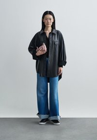 Camisa preta de couro oversized, combinada com jeans azul de perna larga com bainha desfiada e uma pequena carteira rosa. Tênis pretos completam o visual.