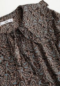 Tissu marron avec des motifs cachemire bleus et beiges ainsi que de petits motifs floraux, rassemblé près d'un décolleté avec une étiquette de vêtement MNG.