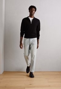 Black zippered sweater, white T-shirt, light blue jeans, and black shoes; model walking on wooden floor against a neutral wall.