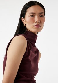 Bordeaux mouwloze coltrui met een gladde textuur, voorzien van een hoge kraag en een aansluitend ontwerp, dat de schouders en de halslijn accentueert.
