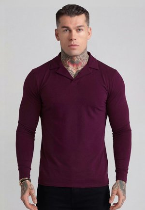 Mężczyzna z tatuażami na szyi i rękach, ubrany w dopasowaną bordową koszulę z długim rękawem, stojący na jednolitym szarym tle.