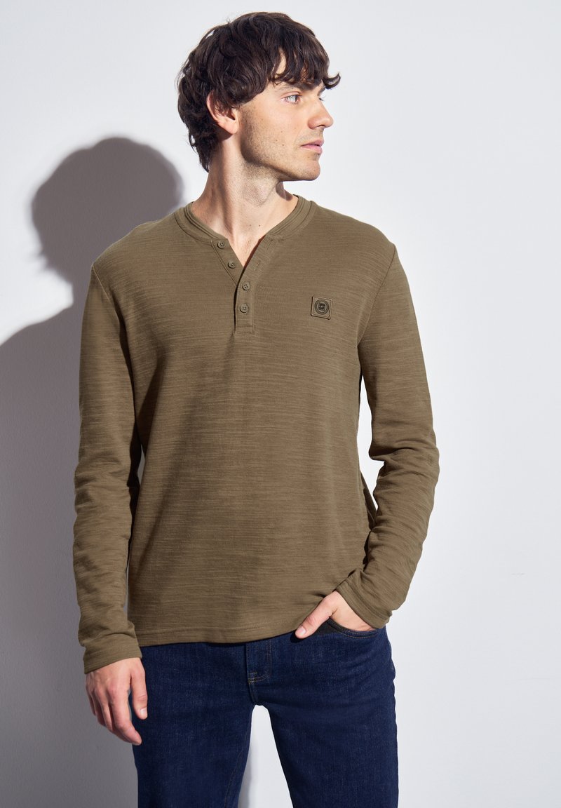 Mörkgrön långärmad henley-tröja med tre knappar, texturerad tyg, rund nederkant och en liten logotyp på vänster bröst.