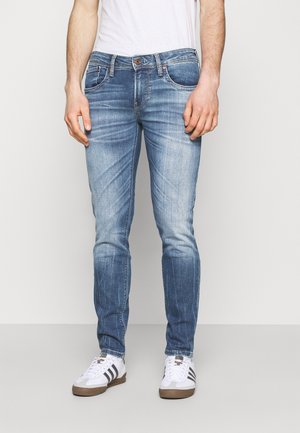 Homme portant un jean slim bleu délavé, un t-shirt blanc et des baskets blanches à bandes noires, debout devant un fond uni.