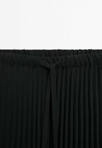 Jupe plissée noire avec une taille élastique et un détail noué ; tissu léger froissé ; les plis verticaux créent de la texture et du mouvement.
