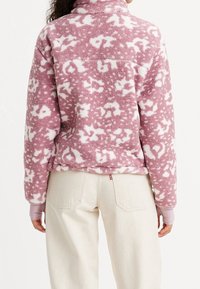 Fleece-Pullover in Pink mit weißem abstraktem Muster, hohem Kragen, langen Ärmeln und weicher Textur. Kombiniert mit hellen Jeans.
