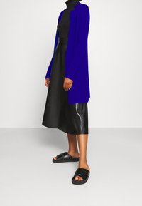 Lång, öppen cardigan i klarblått, tillsammans med en svart kjol i konstläder och svarta sandaler med korsklaffar, vilket visar en lager-på-lager look.