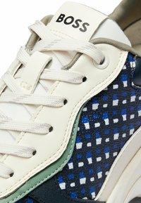 White and navy sneakers with a textured pattern, featuring blue accents, green overlays, and a logo on the tongue. Lace-up closure.