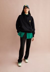 Felpa nera con cappuccio e logo giallo, indossata sopra una maglietta verde e pantaloni neri. Completata con sneakers bianche e verdi e un berretto abbinato.