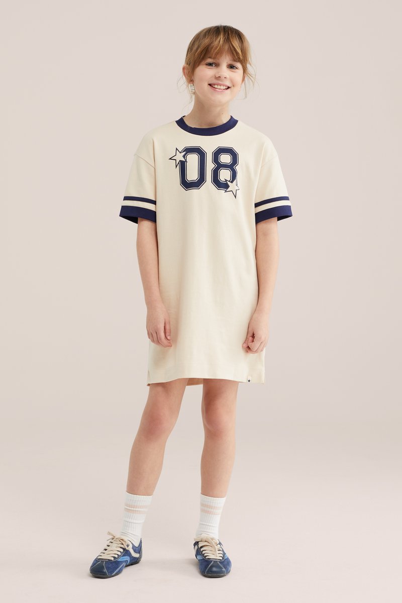 Fille souriante portant une robe t-shirt crème avec le chiffre "08" et des étoiles bleu marine, des manches rayées bleu marine, des chaussettes blanches et des baskets bleu marine, debout sur un fond uni.
