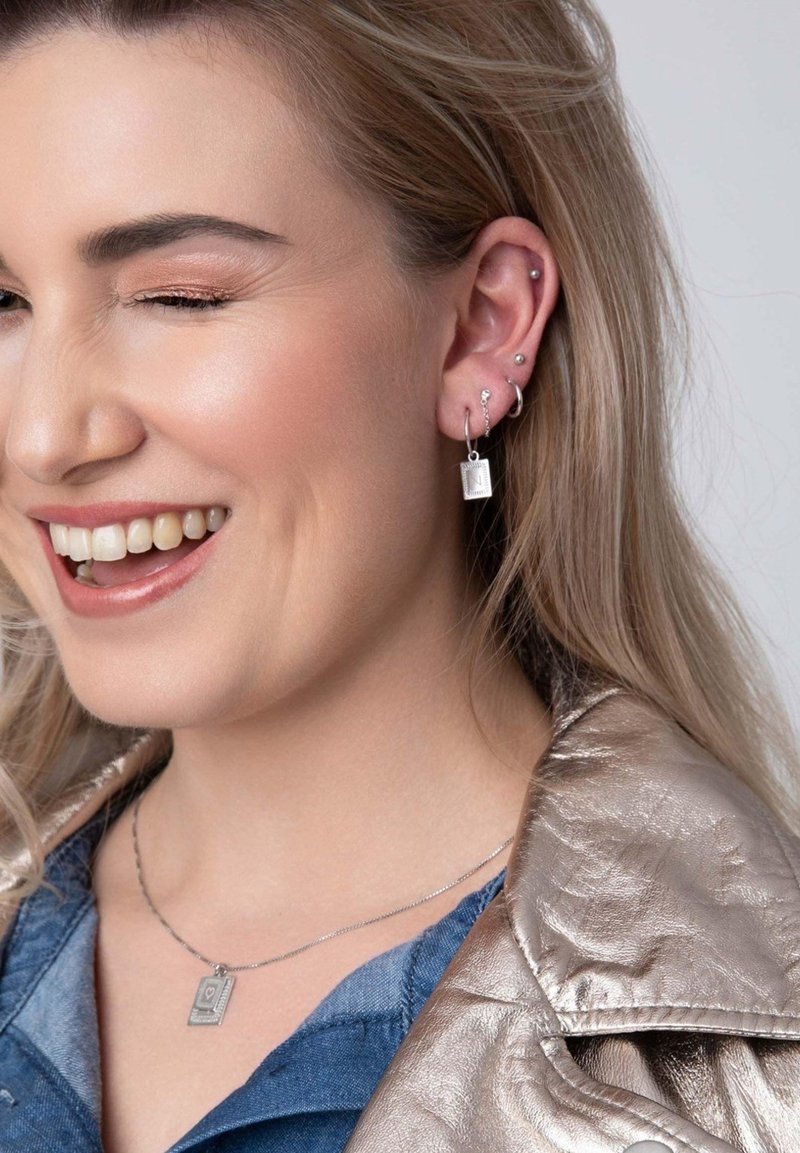 Zilveren oorbellen met een rechthoekige hanger en intrikate patronen, gelaagd met een zilveren ketting, gedragen met een denim shirt en een metallic jack.