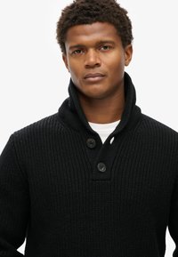 Schwarzer gestrickter Pullover mit hohem Kragen, ausgestattet mit zwei Knöpfen und einem strukturierten Muster. Weicher Stoff mit einem gerippten Design.