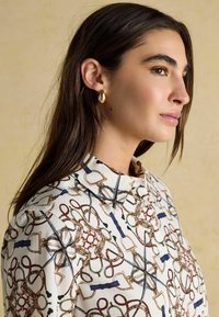 Long-sleeve blouse in a cream fabric with a colorful, abstract print featuring loops and hardware motifs. Side button closure and collar detail.