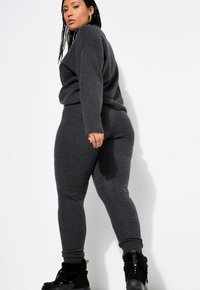 Kohlegrauer Strickpullover und dazu passende schmal geschnittene Hose. Der Stoff ist weich mit einer gerippten Textur an den Bündchen und am Bund. Schwarze Schnürstiefel.
