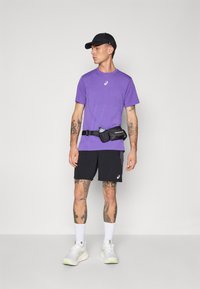 Lila Kurzarmhemd, schwarze Shorts und weiße Sneakers. Verfügt über Tattoos, eine schwarze Gürteltasche und eine schwarze Kappe. Einfaches sportliches Design.