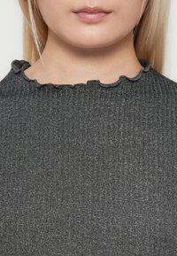 Primo piano del volto inferiore e del collo di una persona che indossa un top grigio scuro a coste con scollatura arricciata.