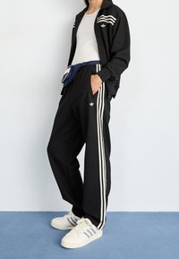 Osoba v černé Adidas sportovní bundě a kalhotách s bílými pruhy, bílém tílku, bílých Adidas teniskách a modré ledvince.