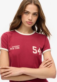 Maronfarvet t-shirt med korte ærmer og cremefarvet kant, der har "SUPERDRY" og "NEW YORK" trykt i hvidt, samt nummeret "54" vist fremtrædende.