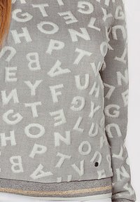 Maglione grigio con lettere bianche in rilievo sparse casualmente sul tessuto, indossato da una persona mostrata dalla parte superiore del torso fino all'anca.