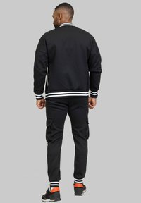 Veste bomber athlétique noire avec un col et des poignets à rayures blanches, associée à un pantalon cargo noir et des baskets noires avec des accents orange.