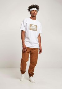 Hvid T-shirt med flerfarvet regnbuegrafik og "SOUTHPOL" tekst, parret med brune tapered bukser og hvide sneakers.