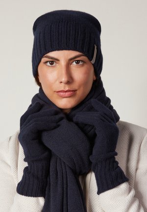 Donna che indossa un cappello, una sciarpa e guanti coordinati in maglia blu scuro, tenendo la sciarpa vicino al corpo su uno sfondo chiaro e neutro.
