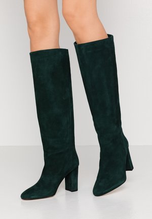 Bottes hautes jusqu'au genou en daim vert foncé avec talons blocs portées sur un fond clair, montrant les vues de face et de côté.