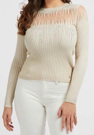 Haut beige à manches longues côtelé avec un haut de poitrine transparent orné de décorations verticales en cristal, porté avec un pantalon blanc.