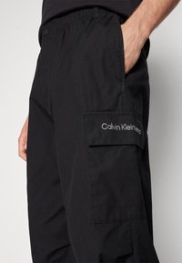 Pantalon cargo noir Calvin Klein avec une poche latérale et une main posée de manière décontractée dans la poche avant, porté avec une chemise noire.