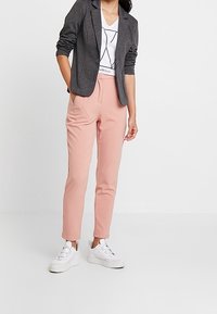 Kvinde iført en grå blazer, hvid grafisk t-shirt, lyserøde bukser og hvide sneakers, stående mod en ensfarvet hvid baggrund.