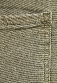 Olivgrön denimtyg med en synlig fickseam och kontrasterande sömnad. Texturen uppvisar ett klassiskt twillmönster.