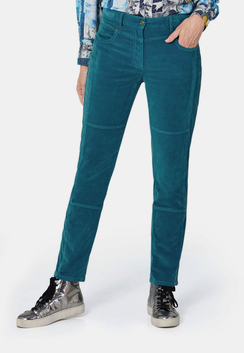 Pantalons en velours côtelé teal au design ajusté, avec des poches frontales et des coutures contrastées. Associés à des baskets montantes métalliques.