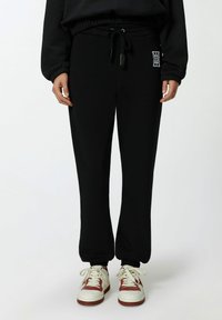 Zwarte joggingsbroek van zacht materiaal, met een tailleband met trekkoord, een taps toelopende pijpdesign en een klein logo aan de linkerkant. Gecombineerd met witte sneakers.