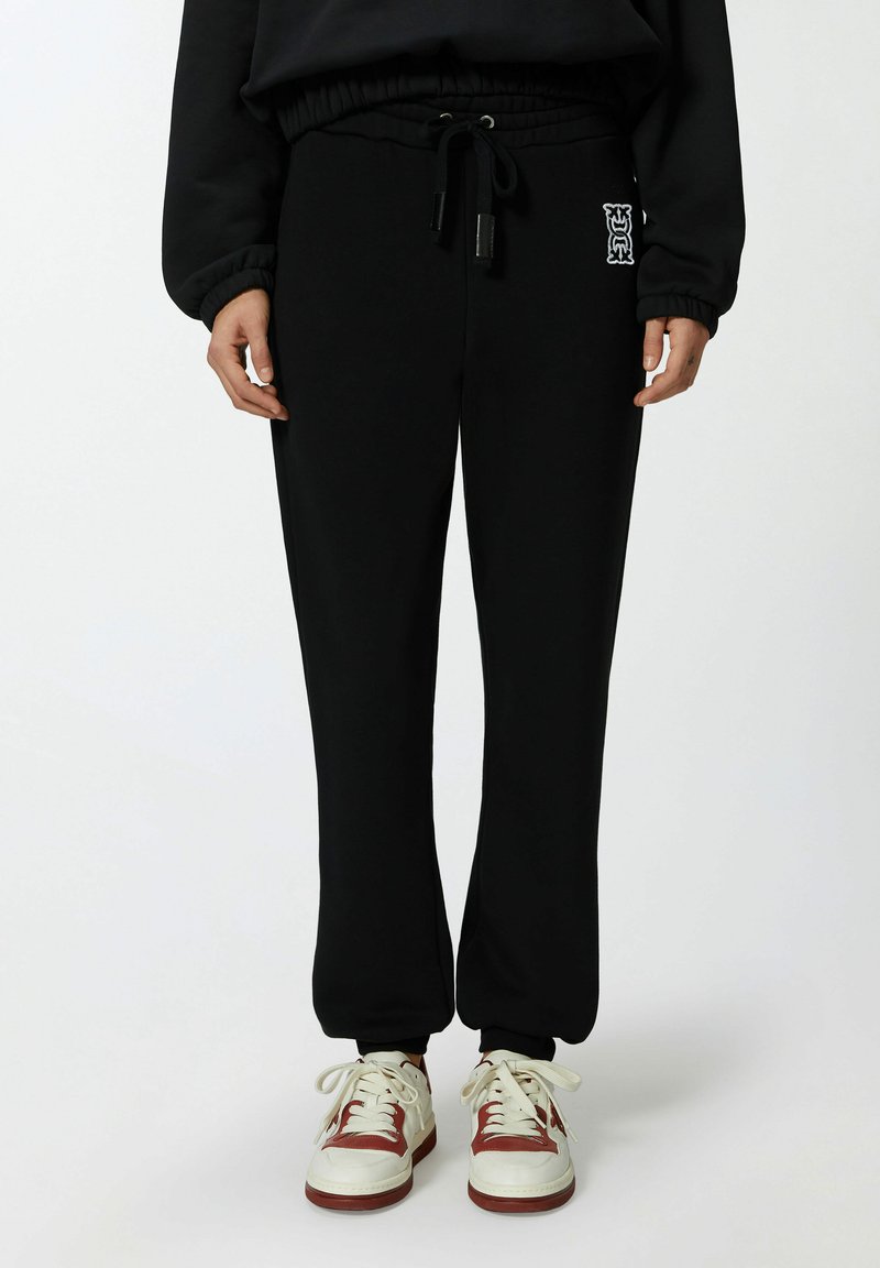 Zwarte joggingsbroek van zacht materiaal, met een tailleband met trekkoord, een taps toelopende pijpdesign en een klein logo aan de linkerkant. Gecombineerd met witte sneakers.