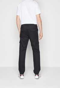 Man in zwarte cargo broeken en witte sneakers, staand met de rug naar de camera tegen een eenvoudige lichte achtergrond.