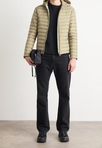 Olivenfarbene Pufferjacke mit Reißverschluss, schwarzer Strickpullover, dunkle Jeans, schwarze Umhängetasche und schwarze Sneakers mit strukturierten Sohlen.