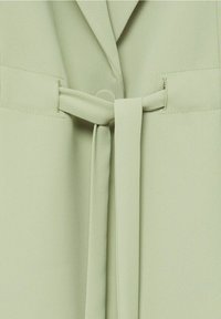 Gros plan sur un vêtement vert clair avec une ceinture en tissu nouée d'un simple nœud au niveau de la taille.