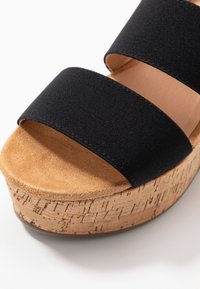 Schwarze Rutschsandale mit zwei Stoffriemen, Kork-Plateau-Sohle und einem strukturierten beigen Fußbett. Die Basis hat ein natürliches Kork-Finish.