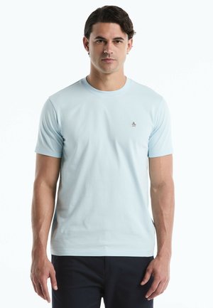 Man draagt lichtblauw T-shirt met korte mouwen en klein pinguïnlogo op de borst, staand tegen een witte achtergrond.