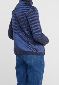 Navy puffer jacket with a quilted pattern, high collar, and shiny finish. Paired with blue denim jeans, showcasing a relaxed fit.