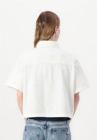 Cropped white shirt with short sleeves, made of lightweight fabric. Features a classic collar and a straight hem, paired with blue denim jeans.