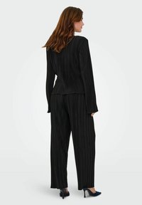 Sort plisseret jakkesæt med en skræddersyet jakke og vide bukser. Stoffet har en struktureret overflade med lodrette striber og lange ærmer.