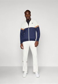 Vīrietis nēsā tumši zilu, baltu un oranžu rāvējslēdzēja jaku, baltas bikses un baltus sporta apavus, stāvot pretī vienkāršam gaišam fonam.