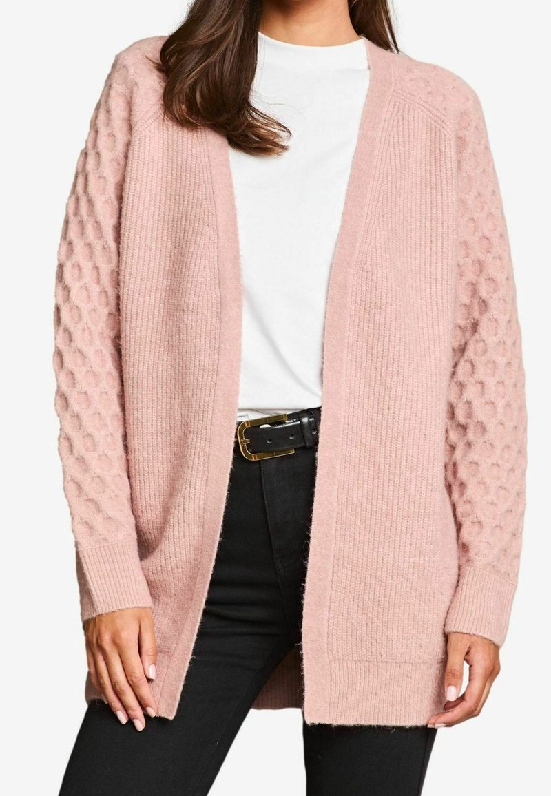 Femme portant un cardigan tricoté texturé rose clair par-dessus une chemise blanche, assorti à un pantalon noir et une ceinture noire avec une boucle dorée.