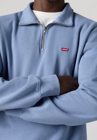 Ljusblå sweatshirt med halvvisselkrage, ribbade ärmslut och en liten röd Levi's-logotyp på bröstet. Mjuk tygtextur.