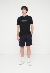 Μαύρο βαμβακερό t-shirt με γραφικό λογότυπο στο μπροστινό μέρος, συνδυασμένο με σκούρο μπλε σορτς και λευκά αθλητικά παπούτσια, τοποθετημένο σε απλό φόντο.